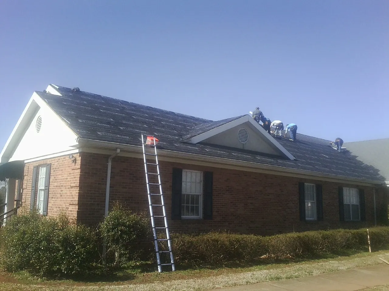 Crew tearing shingles off a brick ranch roof with a ladder set — Metal Roof Repair work by our crew in Buckner